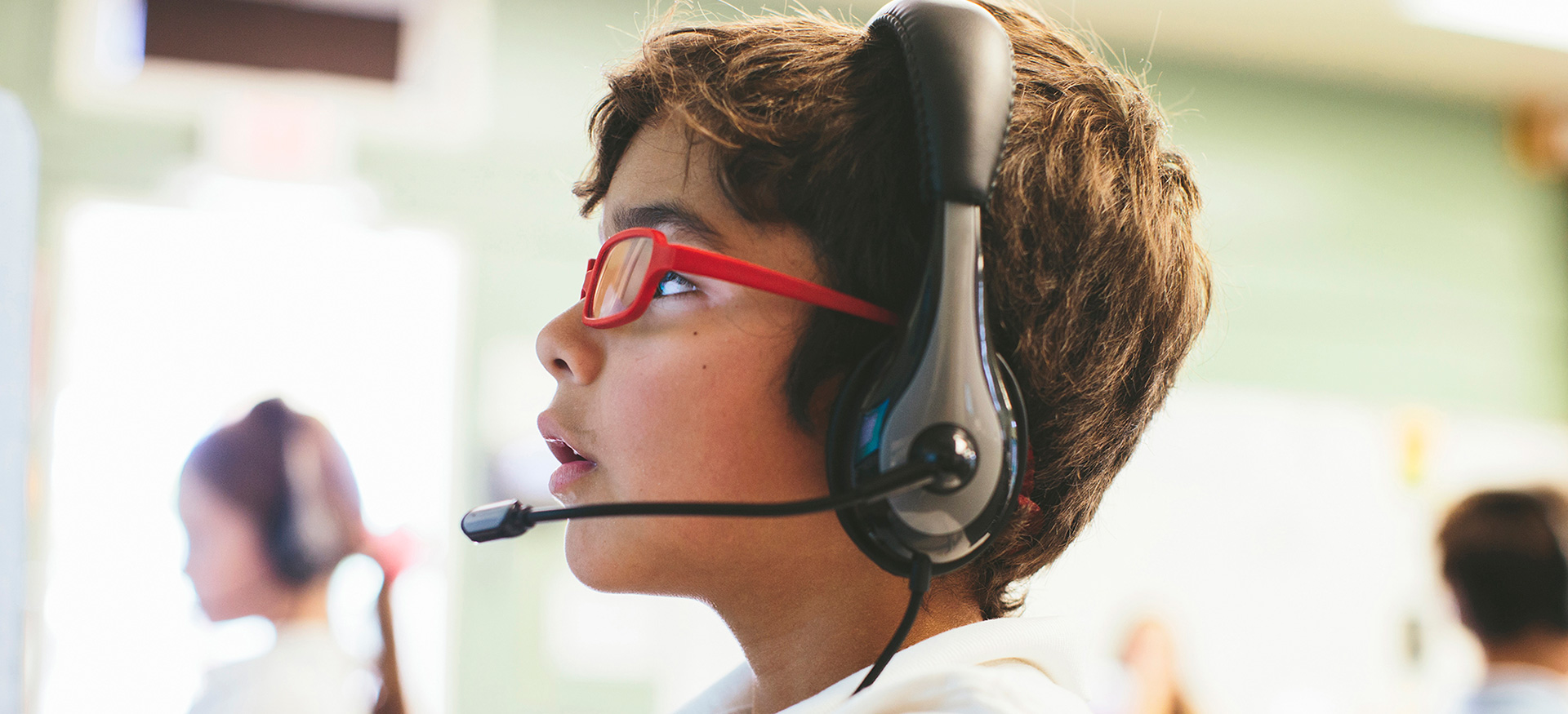 Young student wearing headphones and glasses looking up at a computer.